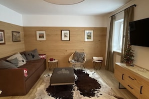 Un salon avec un canapé marron, un petit pouf, un panneau mural en bois, un fauteuil, une petite table, un téléviseur et un sapin de Noël.