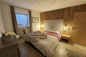 Une chambre avec une tête de lit en bois, un lit recouvert d’un plaid en fourrure, une table de chevet, une fenêtre avec des rideaux et un radiateur.