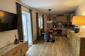 Uma sala de estar moderna com televisão de tela plana, uma área de jantar com mesa e cadeiras, uma cozinha com armários e micro-ondas, e um abajur decorativo.