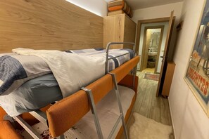 Uma cama beliche com cabeceira de madeira, um guarda-roupa pequeno e uma porta que dá para outro quarto.
