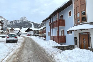 Uma rua coberta de neve com edifícios, um carro e uma montanha ao fundo.