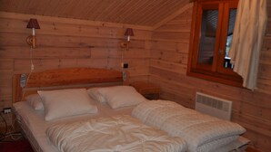 A wooden cabin bedroom with a bed, two bedside lamps, a window with curtains, and a heater.