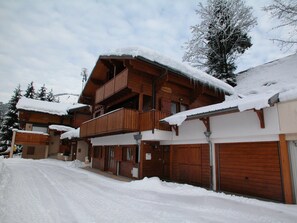 Uma chalé de madeira com varanda, telhado coberto de neve e portões de garagem.