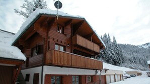 A two-story wooden chalet with a balcony, surrounded by snow-covered trees and a mountain in the background.