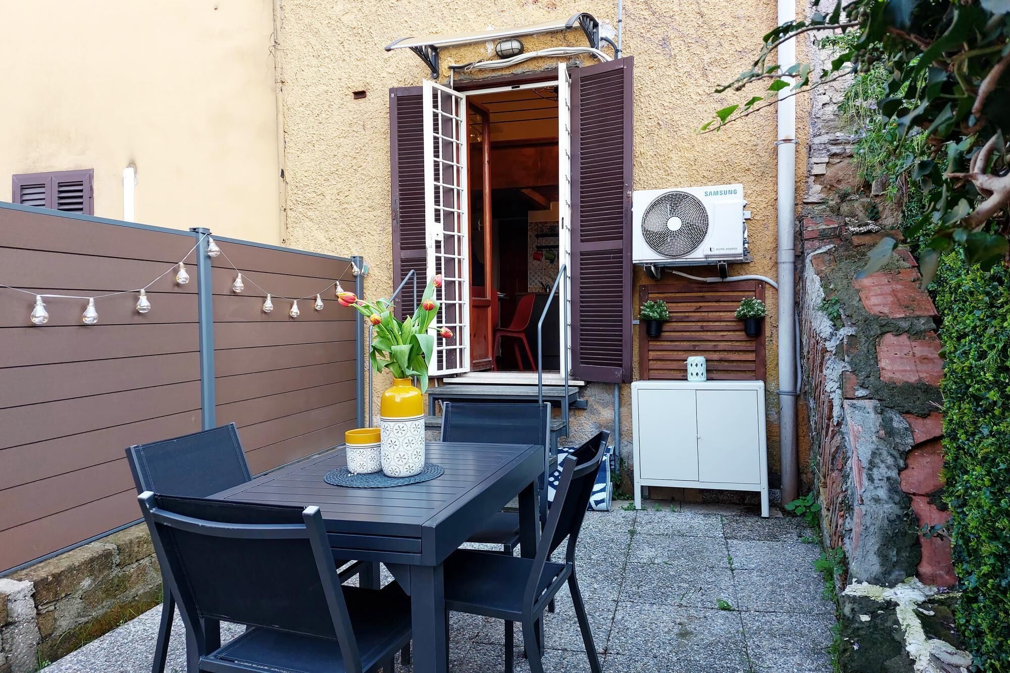 An outdoor patio with a table and chairs, a potted plant, and a wall-mounted air conditioning unit.