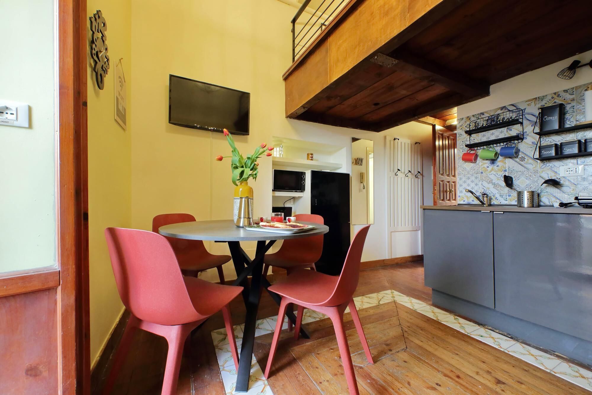 A dining area with a round table and red chairs, a kitchen with a tiled backsplash, and a flat-screen TV mounted on the wall.