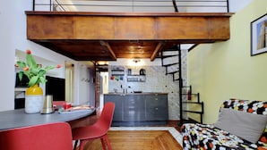 A compact living space with a kitchen area, a dining table with red chairs, and a sofa with a colorful polka dot cover. - 4bnb - Cozy Vatican Loft With Terrace (Rome)