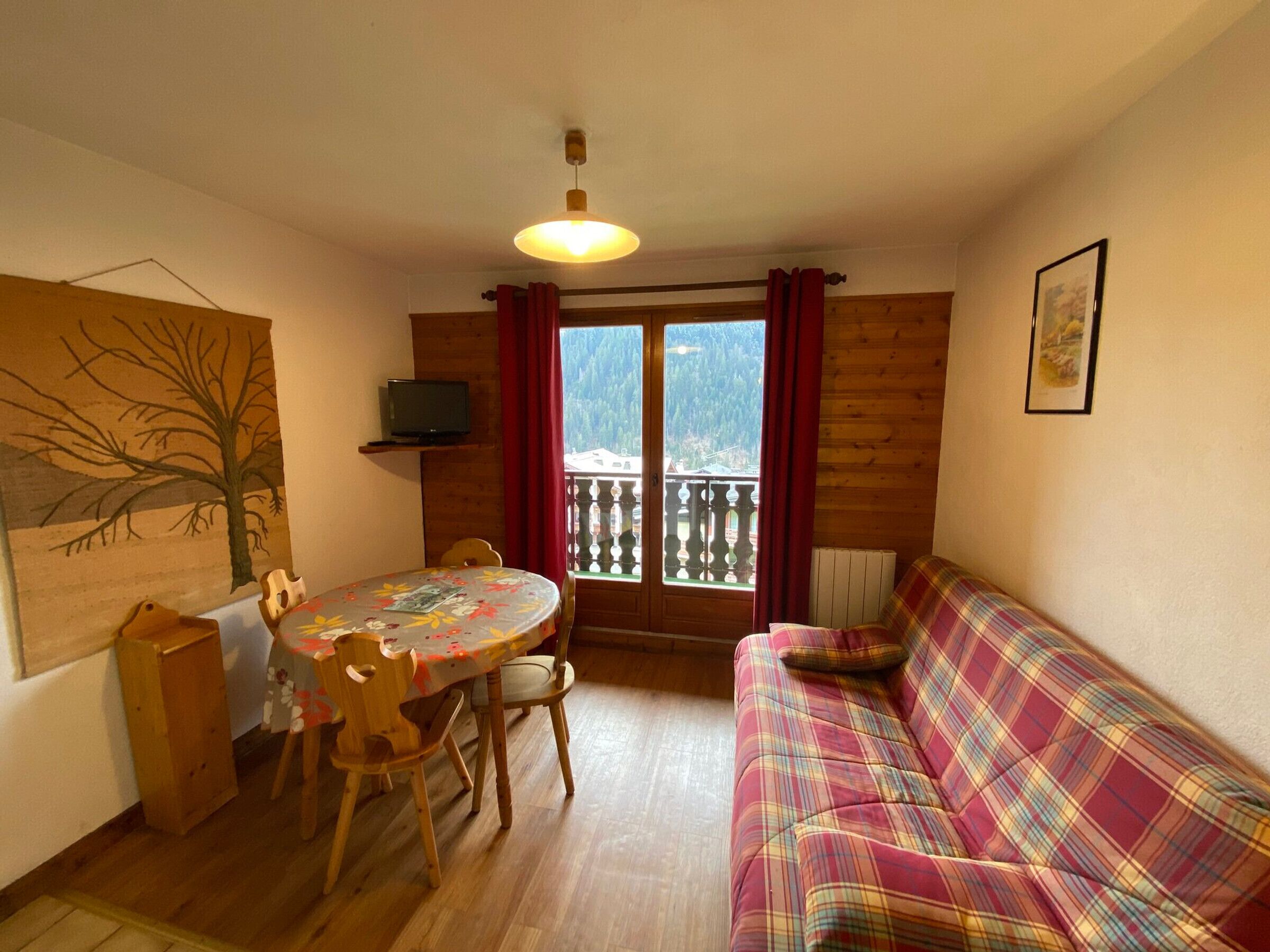 A cozy room with a wooden dining table, chairs, a plaid sofa, a television on a shelf, and a view of mountains through a balcony door.