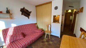 Una sala de estar con un sofá rojo estampado, una mesa de comedor de madera, una silla de madera, un taburete pequeño de madera, un reloj en la pared, un estante de madera con una botella y una puerta que da a una cocina.