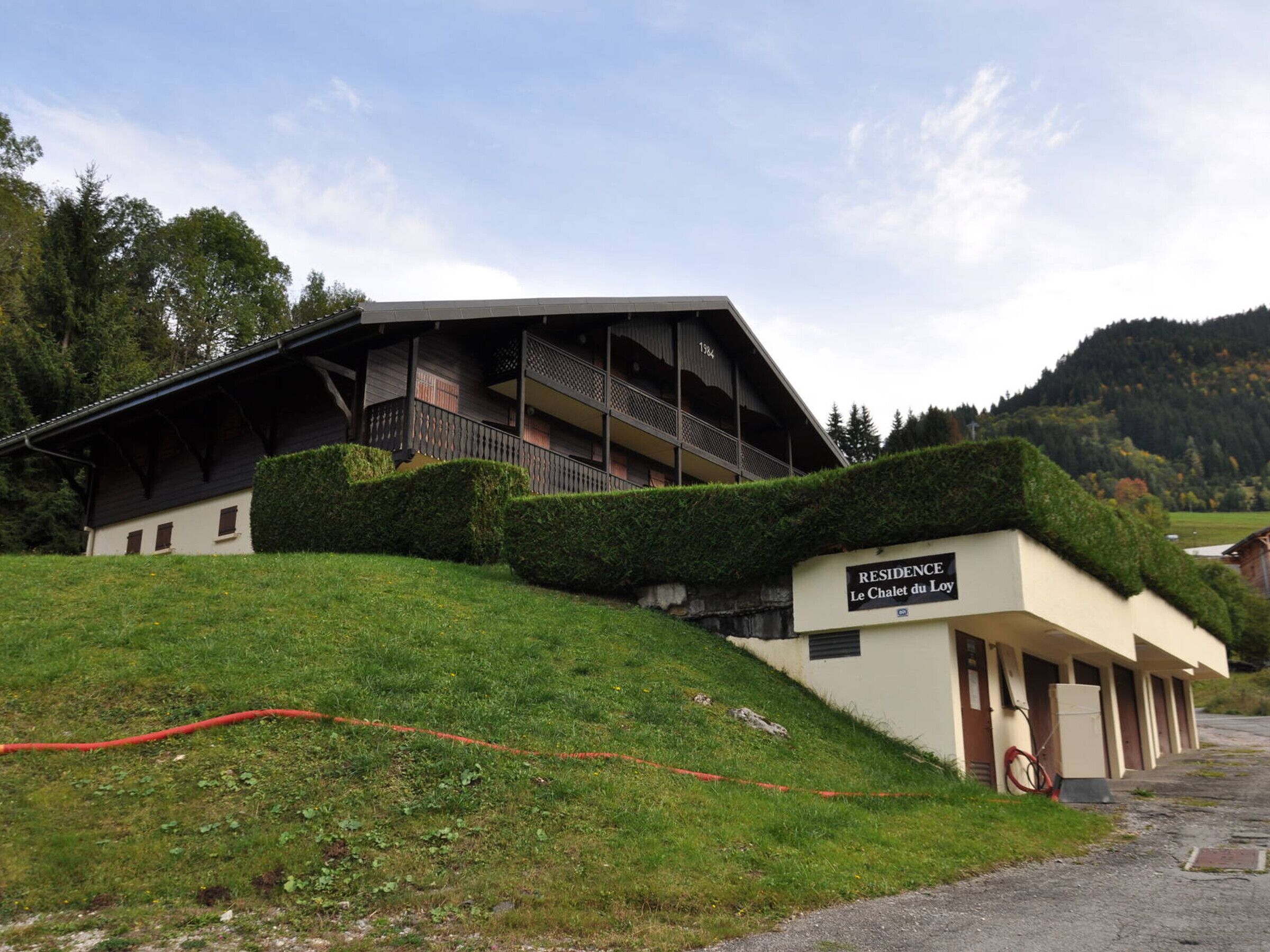 A building with a sign reading "RESIDENCE Le Chalet du Loy". It features a multi-story structure with balconies, a sloped roof, and a surrounding hedge.