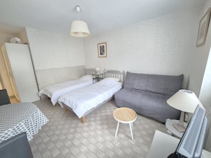 Une petite chambre à coucher, bien rangée, avec deux lits simples, un canapé gris, une petite table ronde et un bureau avec un ordinateur.