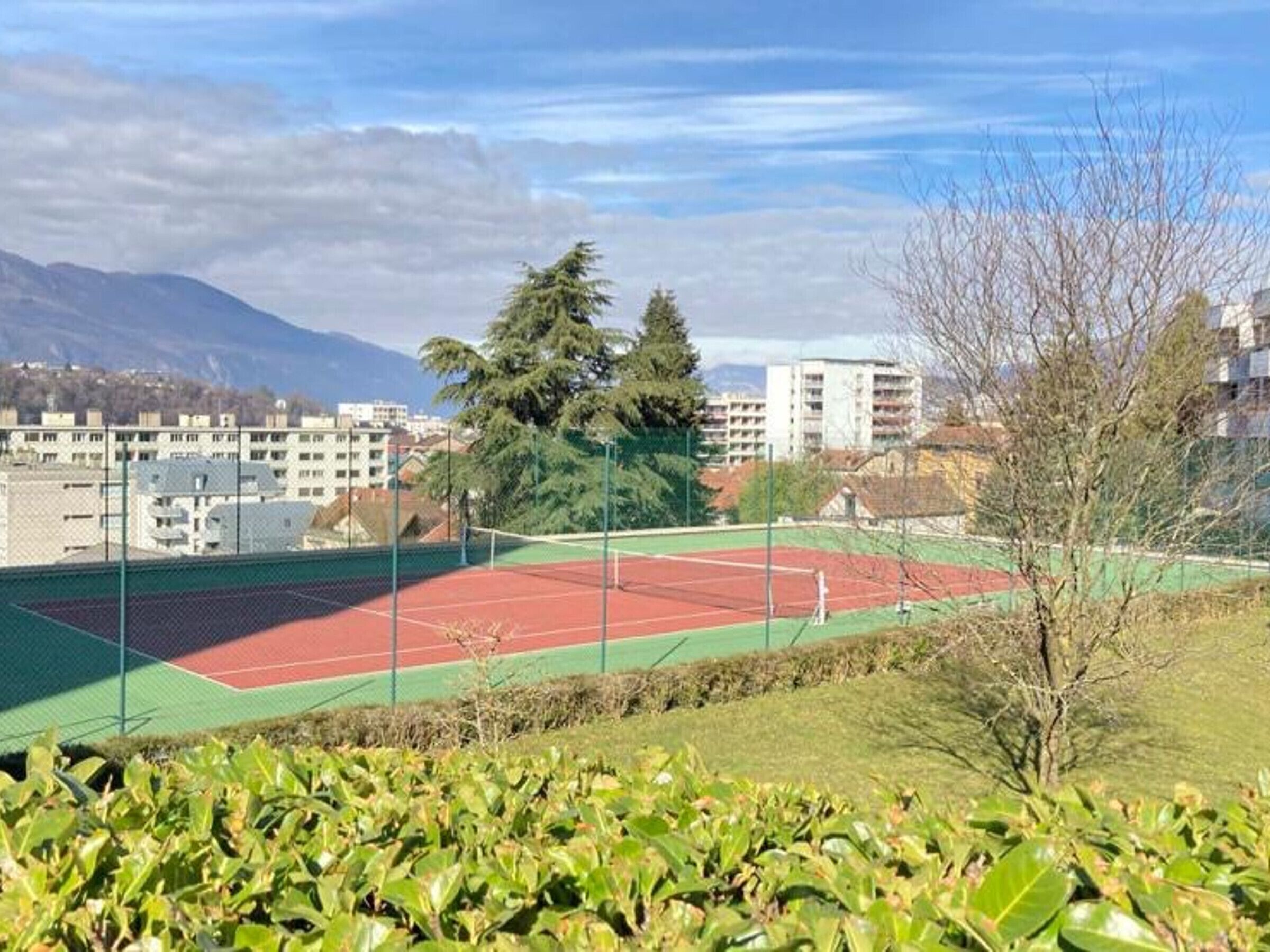 A tennis court with a green and red surface, surrounded by greenery and buildings in the background.
