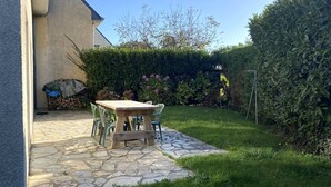 Eine Steinterrasse mit einem Holztisch und Stühlen, einer Hecke und einem Gebäude im Hintergrund.