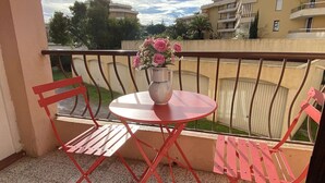 Un balcon avec une petite table ronde et deux chaises rouges, un vase rempli de fleurs roses, et une vue sur des bâtiments en arrière-plan.