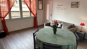 Un coin repas avec une table ronde, des chaises, un vase de fleurs, un canapé, une lampe et un mur orné d’une inscription de bienvenue.
