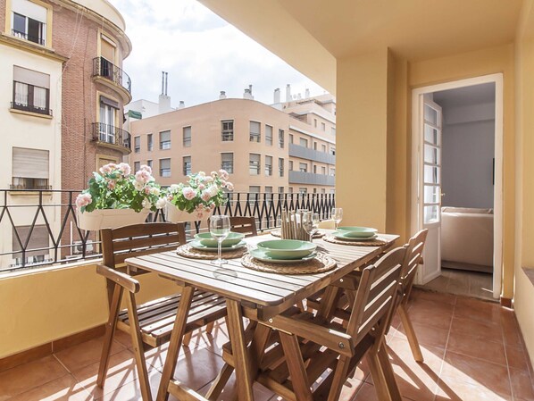 A balcony with a wooden table set for dining, potted plants, and a view of surrounding buildings.
