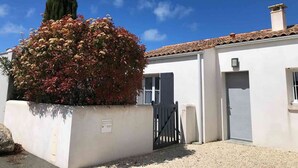 A white building with a tiled roof, a gated entrance, and a small garden with a tree and shrubs.