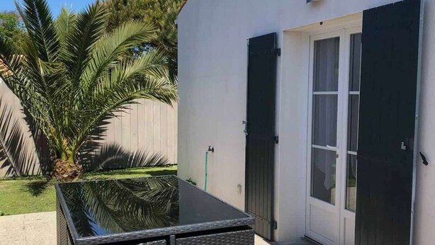 A modern outdoor patio with a glass-top table, a door with black shutters, and a palm tree in the background.