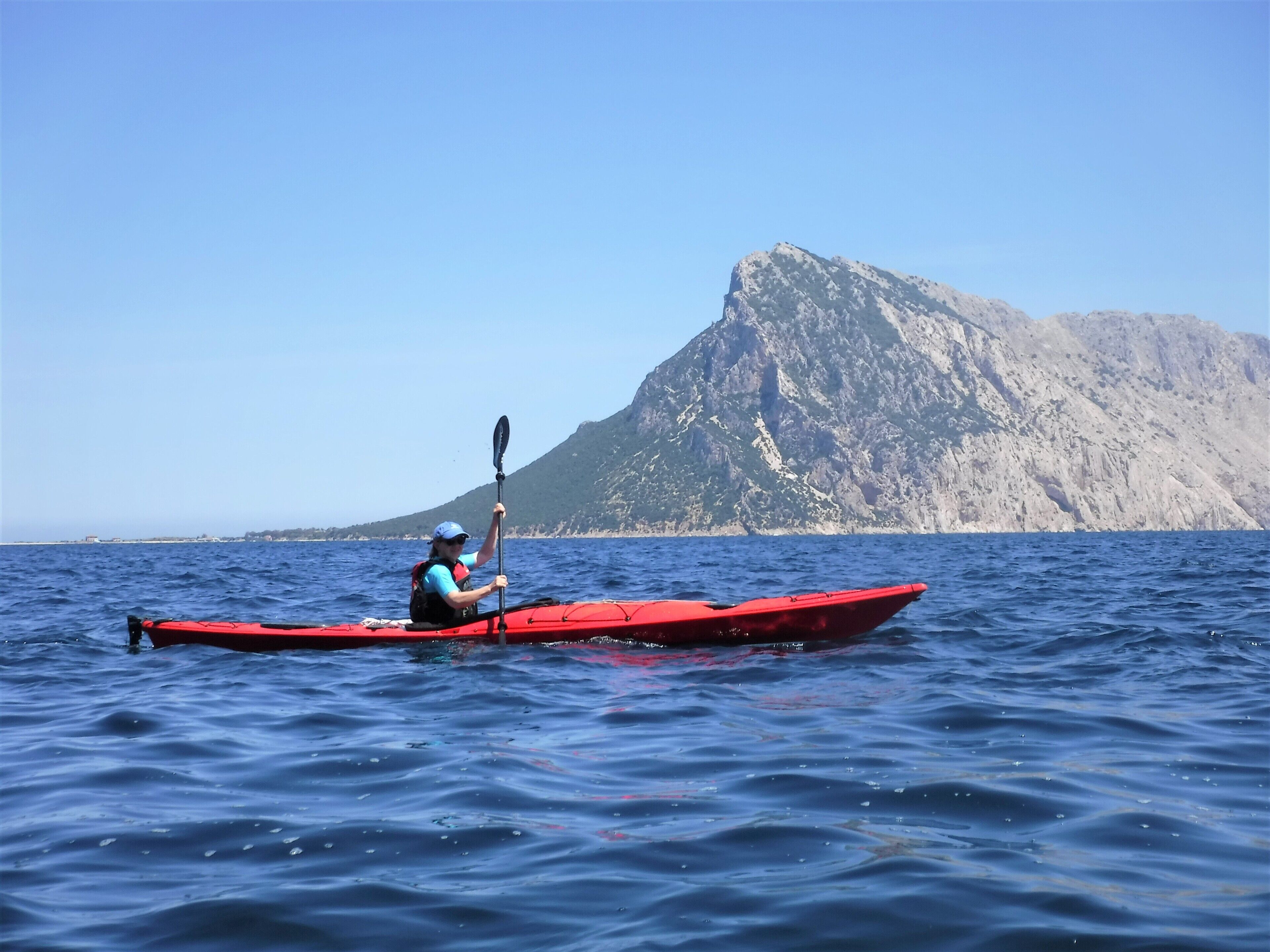 A person kayaking on a calm sea with a mountain in the background.