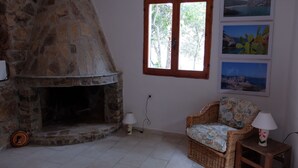 A stone fireplace, a wicker chair with a floral cushion, and a window with a view of trees.