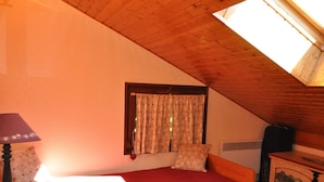 A bedroom with a sloped ceiling, a single bed with a red mattress, a nightstand with a lamp, a dresser with a decorative panel, and a window with floral curtains. - Charming apartment near center and cable car with parking (Châtel)