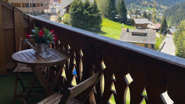 En træterrasse med et bord og stole, med udsigt over en bjerglandsby med huse og grønt.