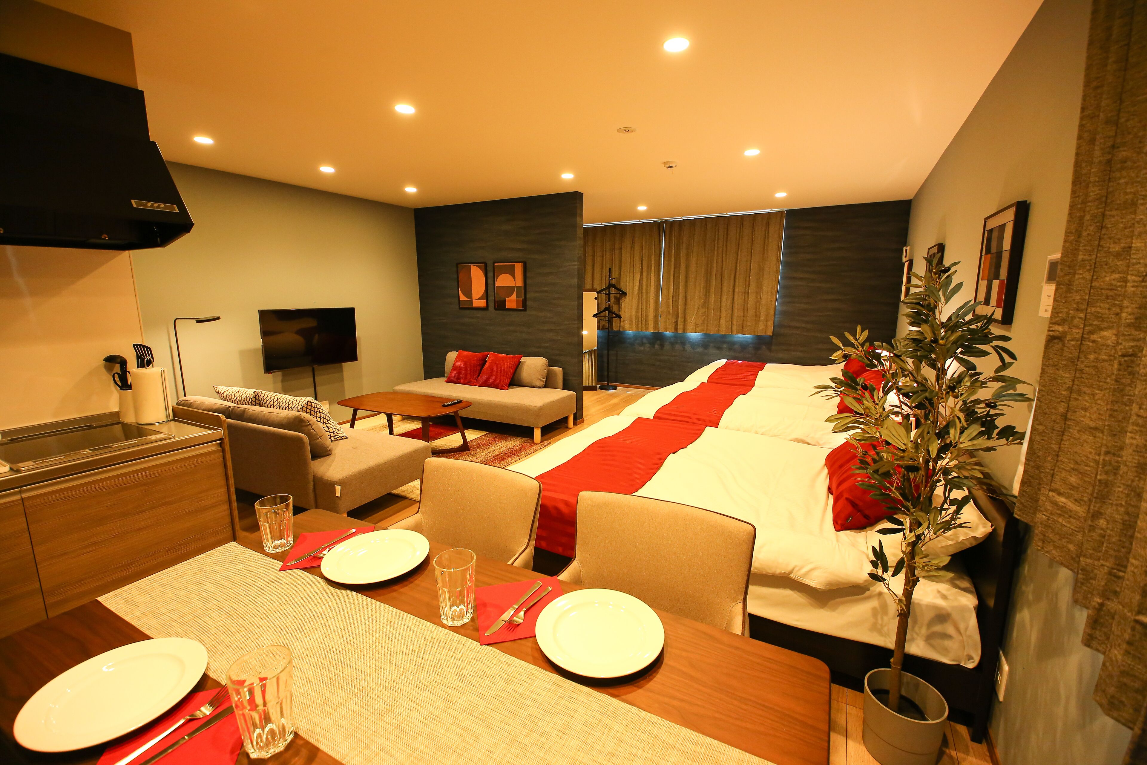 A hotel room with a dining area, a sofa, a bed with a red blanket, a TV, and a plant.