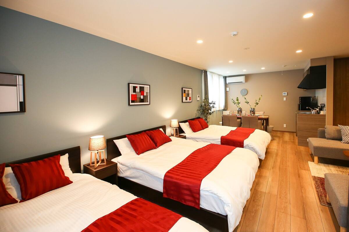Quarto de hotel com três camas, área de refeições e cozinha compacta.