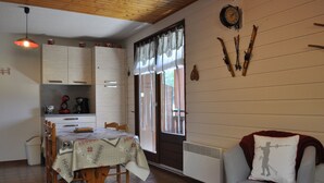 Un comedor con una mesa dispuesta para cuatro, una silla con cojín, una cocina al fondo y una ventana con cortinas.