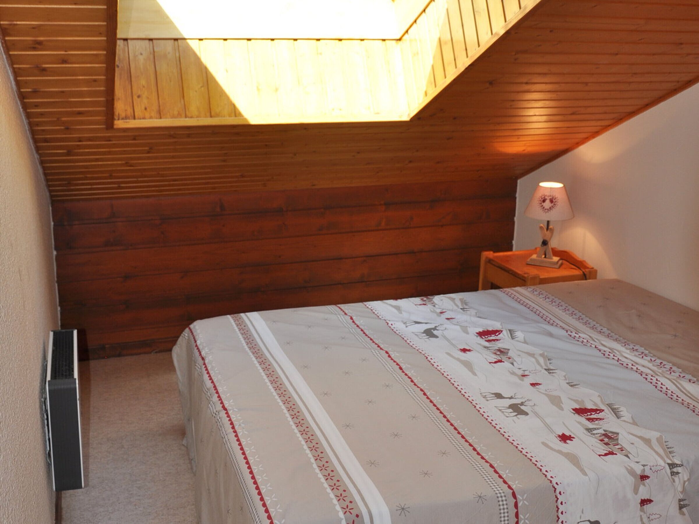 Una camera da letto con soffitto spiovente, pareti in legno, un letto con copriletto floreale, un comodino con una lampada e un termosifone.