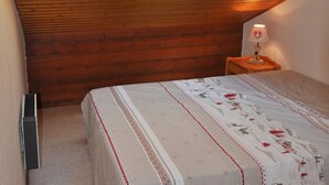 Un dormitorio con techo inclinado, paredes de madera, una cama con colcha estampada, una mesita de noche con lámpara y un radiador.