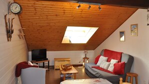 Una habitación acogedora en el ático, con techo inclinado de madera, un sofá con cojines rojos, un televisor, una mesita y un reloj en la pared.