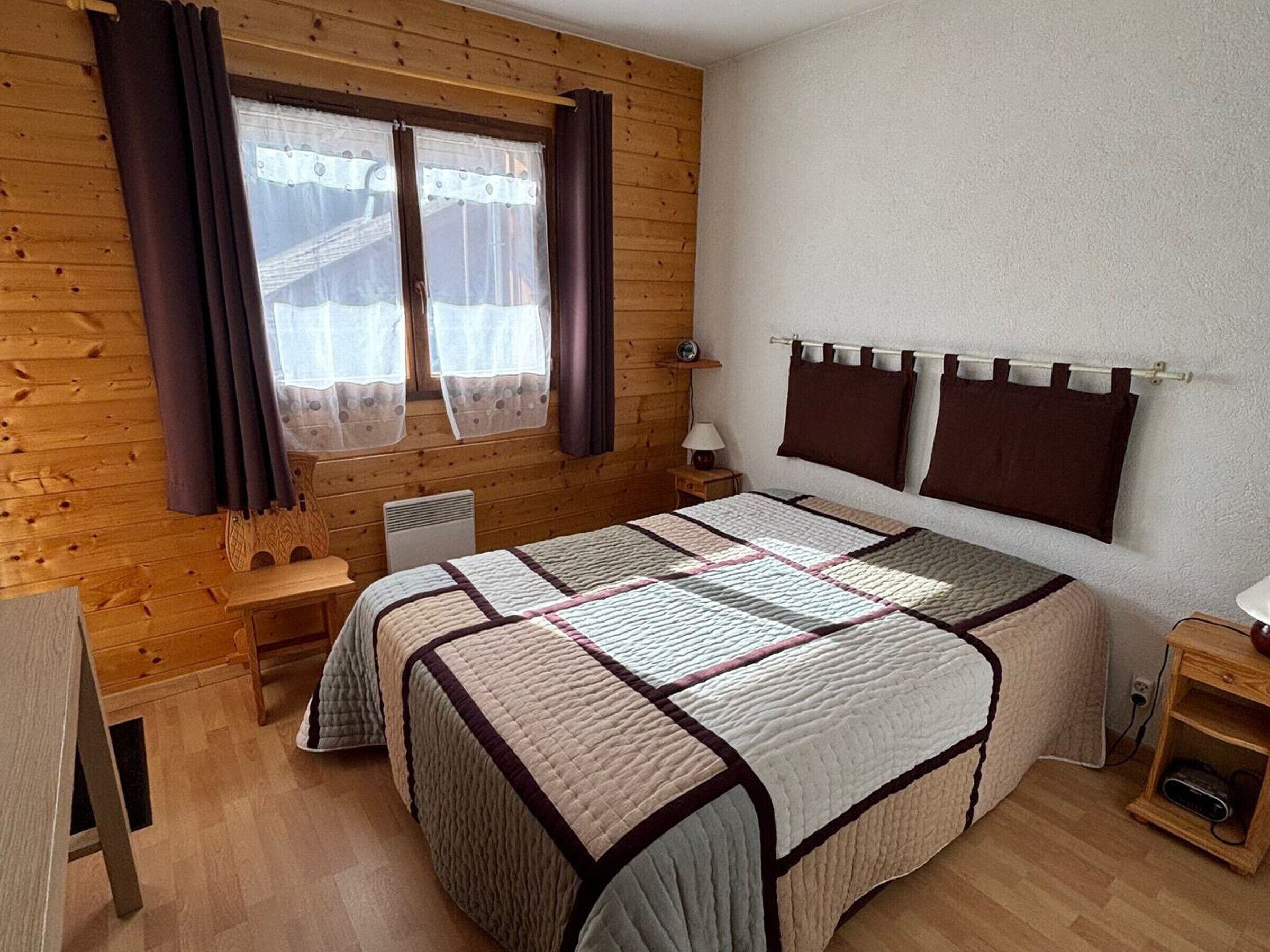 Una habitación con paneles de madera, una cama, una ventana con cortinas, una mesita de noche y un buró.
