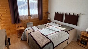 Ein Zimmer mit Holzwänden, einem Bett, einem Fenster mit Vorhängen, einem Nachttisch und einem Beistelltisch.