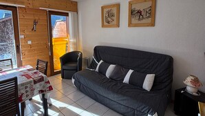 Ein gemütliches Zimmer mit einem grauen Sofa, einem kleinen Tisch mit rotem und weißem Tischläufer, zwei gerahmten Bildern an der Wand und einer Glas-Schiebetür, die auf einen Balkon führt.
