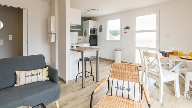 Ein modernes Wohnzimmer mit einem grauen Sofa, einem Korbstuhl und einem Esstisch mit Stühlen. Im Hintergrund ist eine Küche mit weißen Schränken und einem Fenster zu sehen.