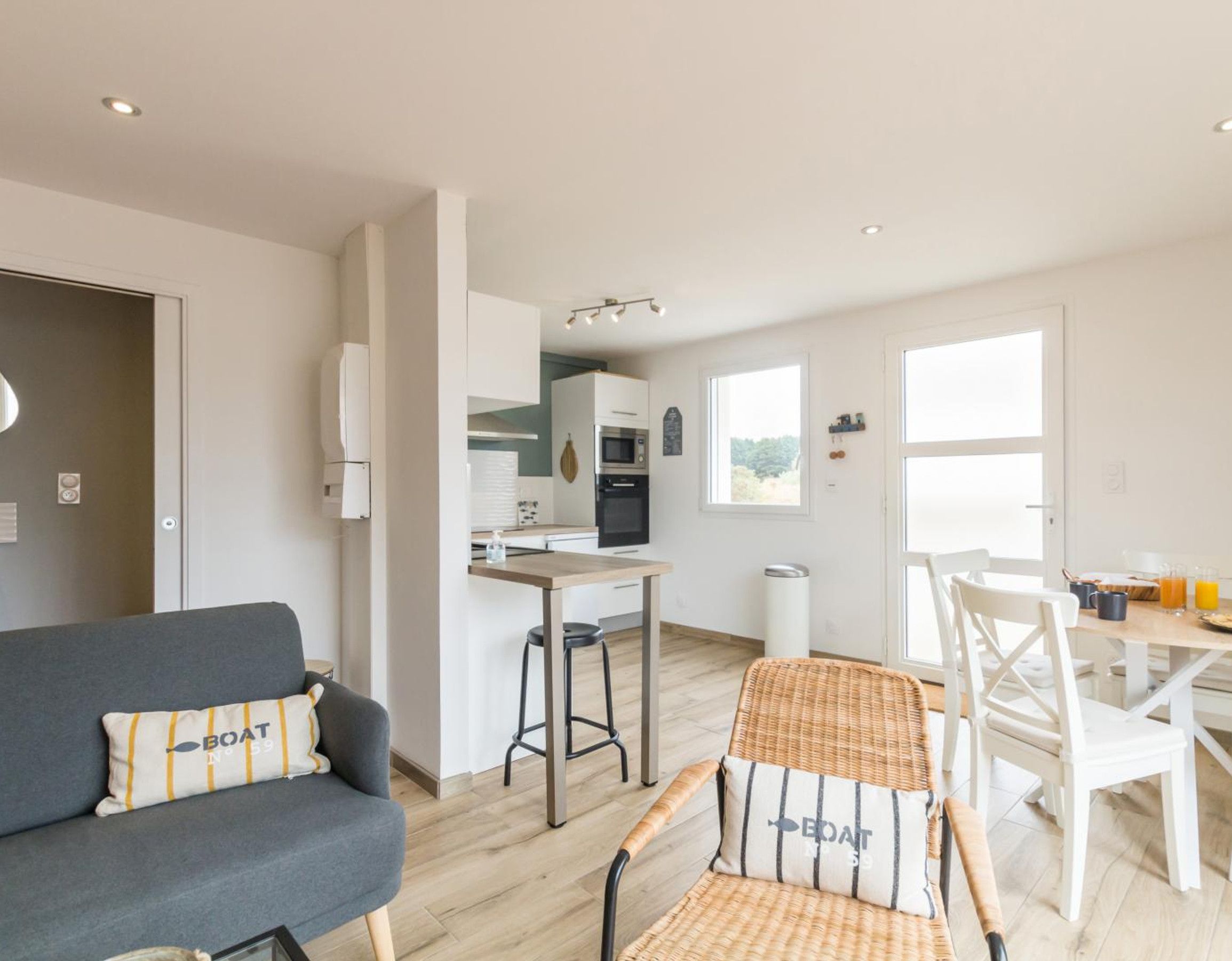 Una sala moderna con un sofá gris, un sillón de mimbre y una mesa de comedor con sillas. Se ve una cocina al fondo con armarios blancos y una ventana.