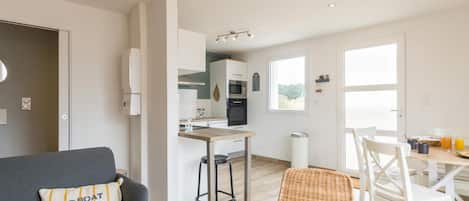 Una sala moderna con un sofá gris, un sillón de mimbre y una mesa de comedor con sillas. Se ve una cocina al fondo con armarios blancos y una ventana.