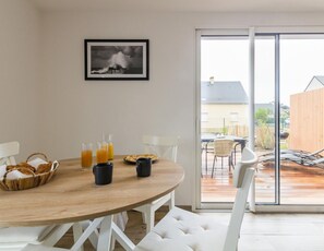 Un comedor con mesa de madera, sillas blancas y puerta de vidrio que da a un patio exterior.