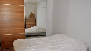 Un dormitorio con un armario de madera, una cama con ropa de cama blanca, una mesita de noche y un espejo que refleja la habitación.