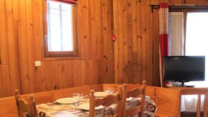 Una sala menjador de fusta amb una taula posta per a quatre, una televisió sobre un suport i una finestra amb cortines.
