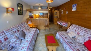 A cozy living room with two patterned sofas, a wooden coffee table, and a kitchen area with wooden cabinets and a hanging light fixture. - Cozy studio with balcony and WIFI near Linga slopes (Châtel)