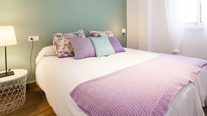 A neatly made bed with a white comforter and a purple knitted throw. There is a floral pillow set on the bed, and a white air conditioning unit mounted on the wall above it.