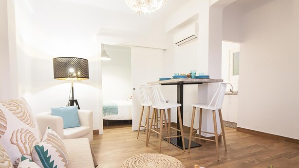 A modern living room with a sofa, a dining table with chairs, a lamp, and a small kitchen area.