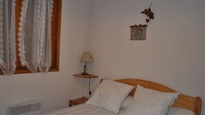A bedroom with a wooden bed, white bedding, a bedside lamp, a window with curtains, a wall-mounted picture, and a radiator. - Cozy apartment near slopes with balcony and parking (Châtel)