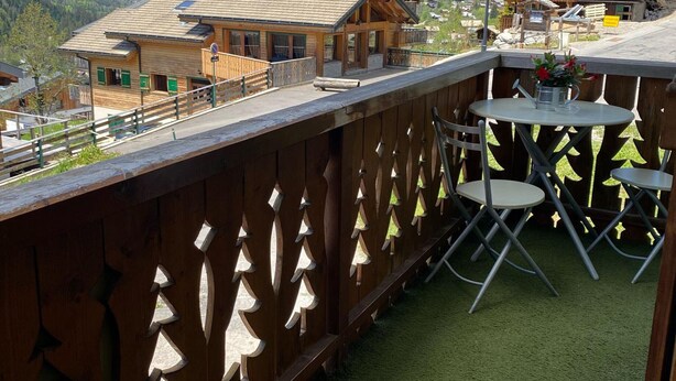 Un balcón de madera con vistas a una cordillera, una pequeña mesa con sillas y una valla de madera con adornos decorativos.