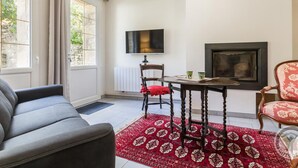 Un salon avec un canapé gris, un tapis rouge à motifs, une table en bois avec des chaises, un téléviseur fixé au mur et une cheminée.