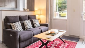 Un salon avec un canapé, des coussins à motifs, une petite table sur laquelle se trouve une assiette de nourriture, une lampe et une vue sur l’extérieur par une porte.