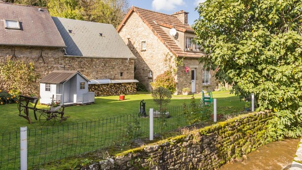 Un bâtiment en pierre avec un petit abri attenant, une cour herbeuse et un muret menant à un ruisseau.