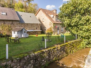 Un bâtiment en pierre avec un petit abri attenant, une cour herbeuse et un muret menant à un ruisseau.